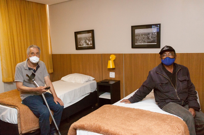 dois homens idosos com deficiência sentados em duas camas de um quarto de hotel. 